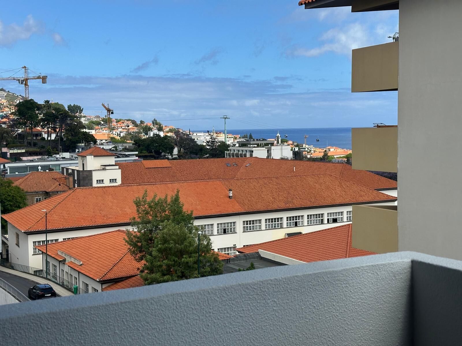 A coastal cityscape with terracotta-roofed buildings, a clear sky, and a view of the sea.
