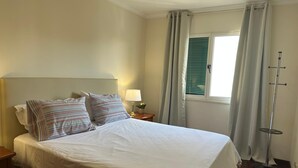A neatly made bed with white linens and striped pillows, a bedside lamp, a window with curtains, and a floor lamp.