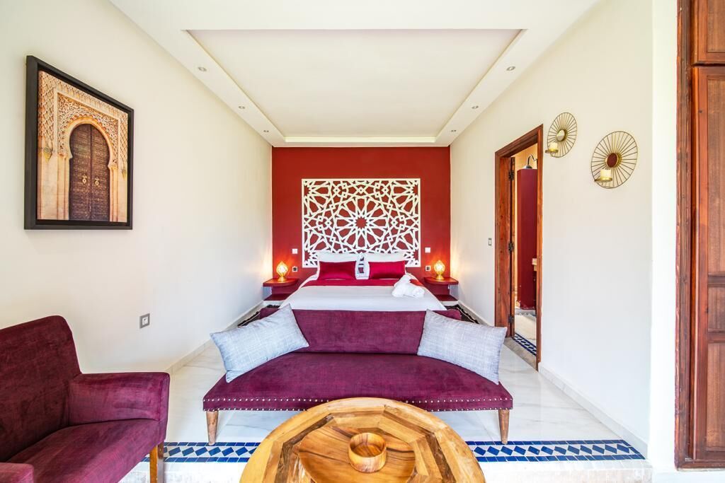 A bedroom with a large bed, a red accent wall, a decorative wall piece, and a wooden coffee table.