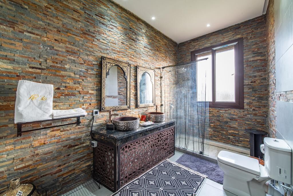 A bathroom with a stone wall, a glass shower, a sink with two bowls, and a toilet.