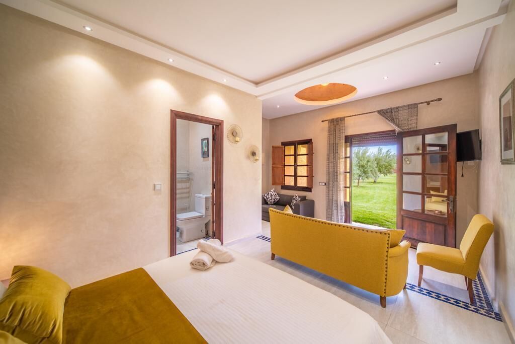 A bedroom with a bed, a yellow sofa, a chair, a bathroom, and a view of the outdoors.