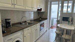A modern kitchen with white cabinetry, a built-in washing machine, a coffee maker, and a dining area with a table and chairs. - Robert Apartment in Ajuda (Funchal)