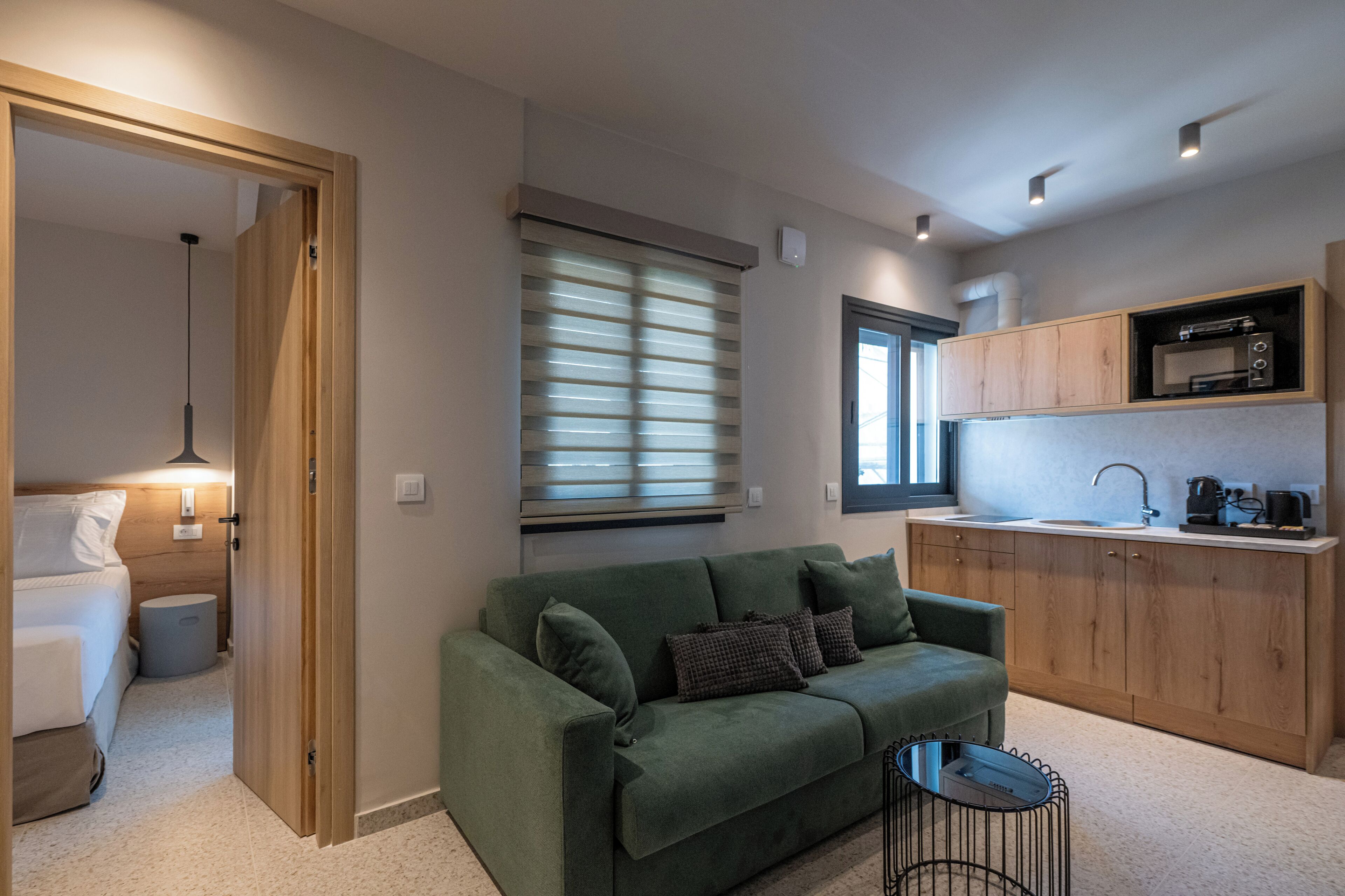 Een ruimtelijk ingerichte leefruimte met een groene bank, een klein rond tafeltje, een kitchenette met een magnetron, en een raam met jaloezieën.