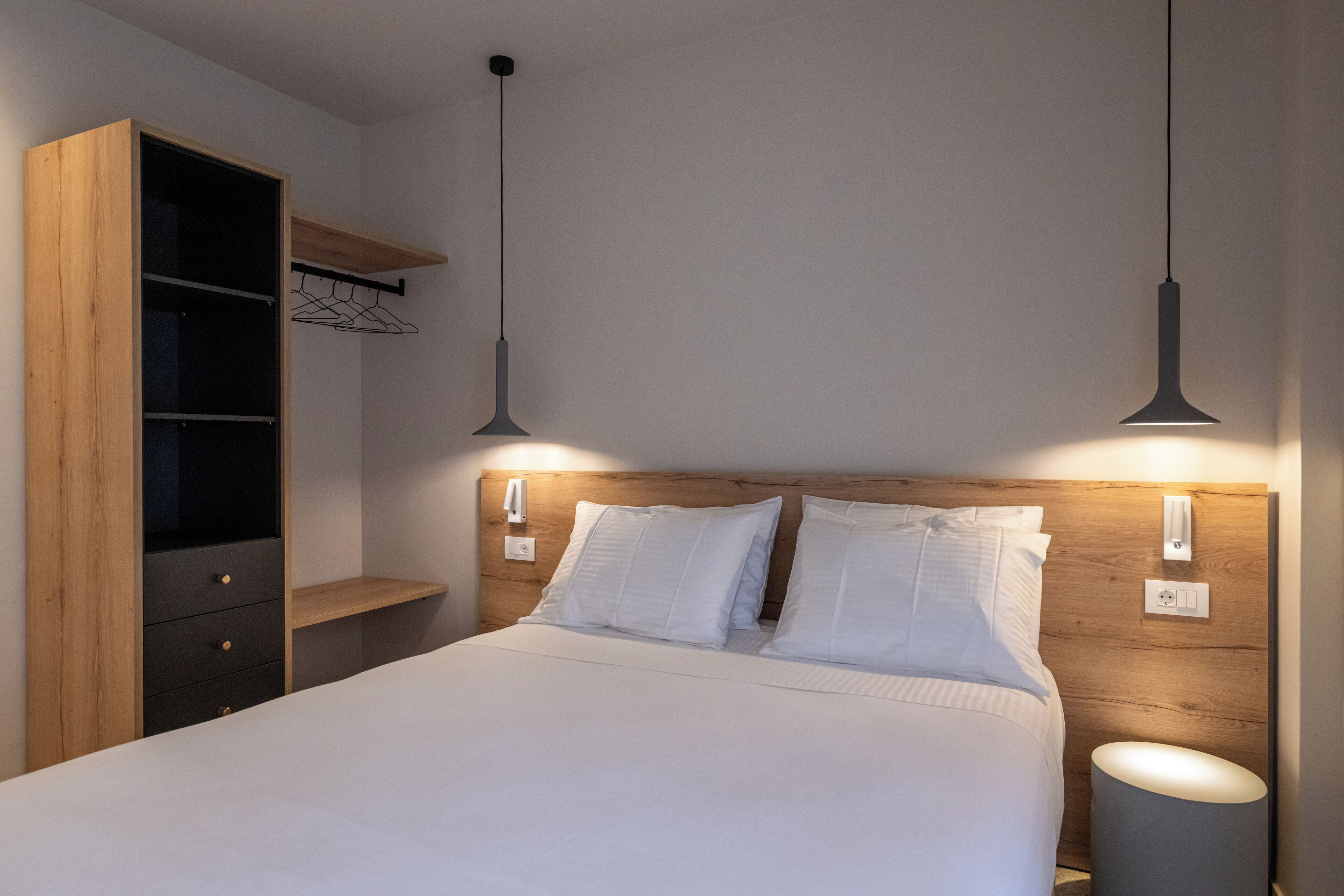 A bedroom with a wooden headboard, a bed with white linens, two pendant lights, and a wardrobe with a hanging rack.