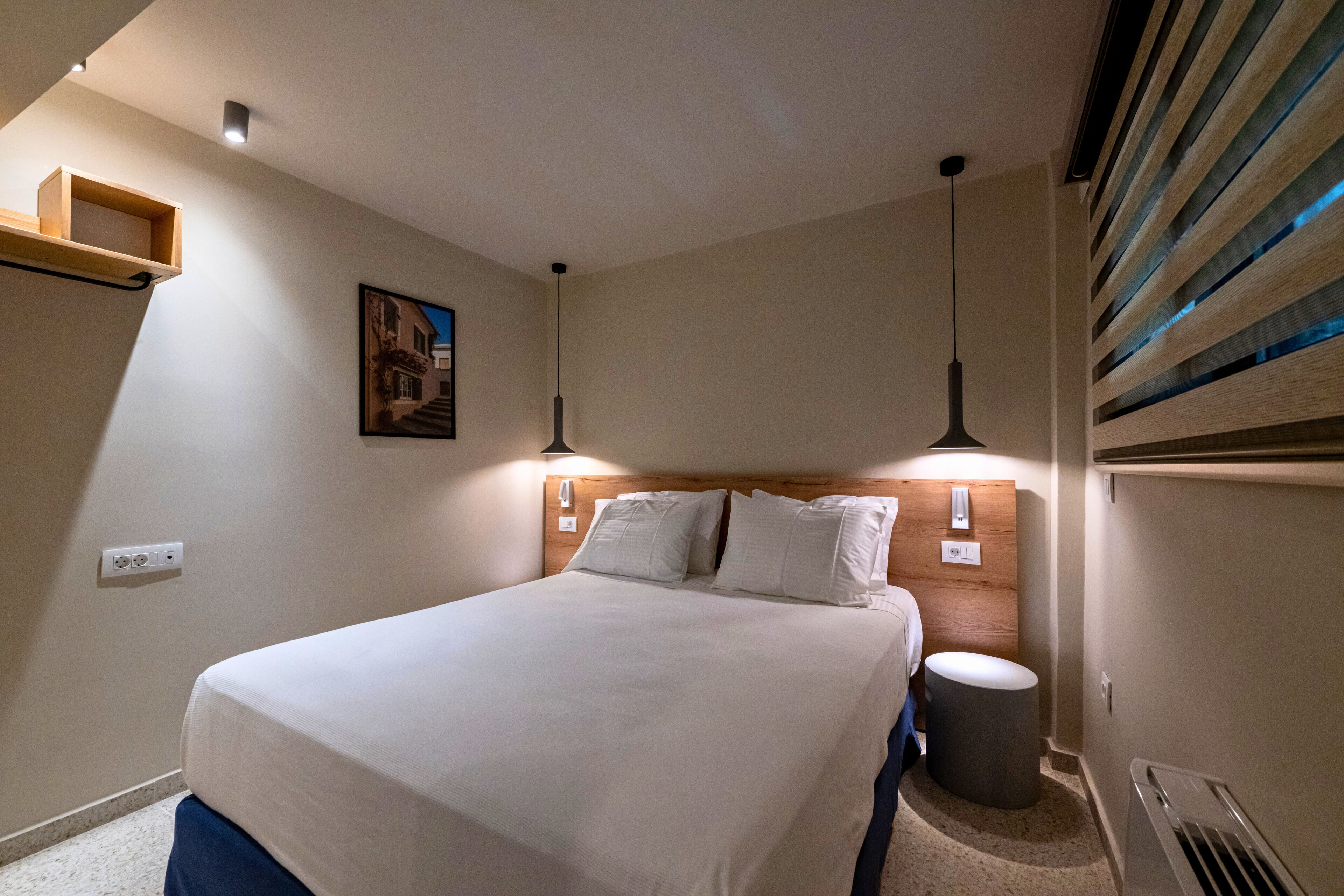 A hotel room with a bed, bedside lamps, a small trash bin, and a wall-mounted shelf.