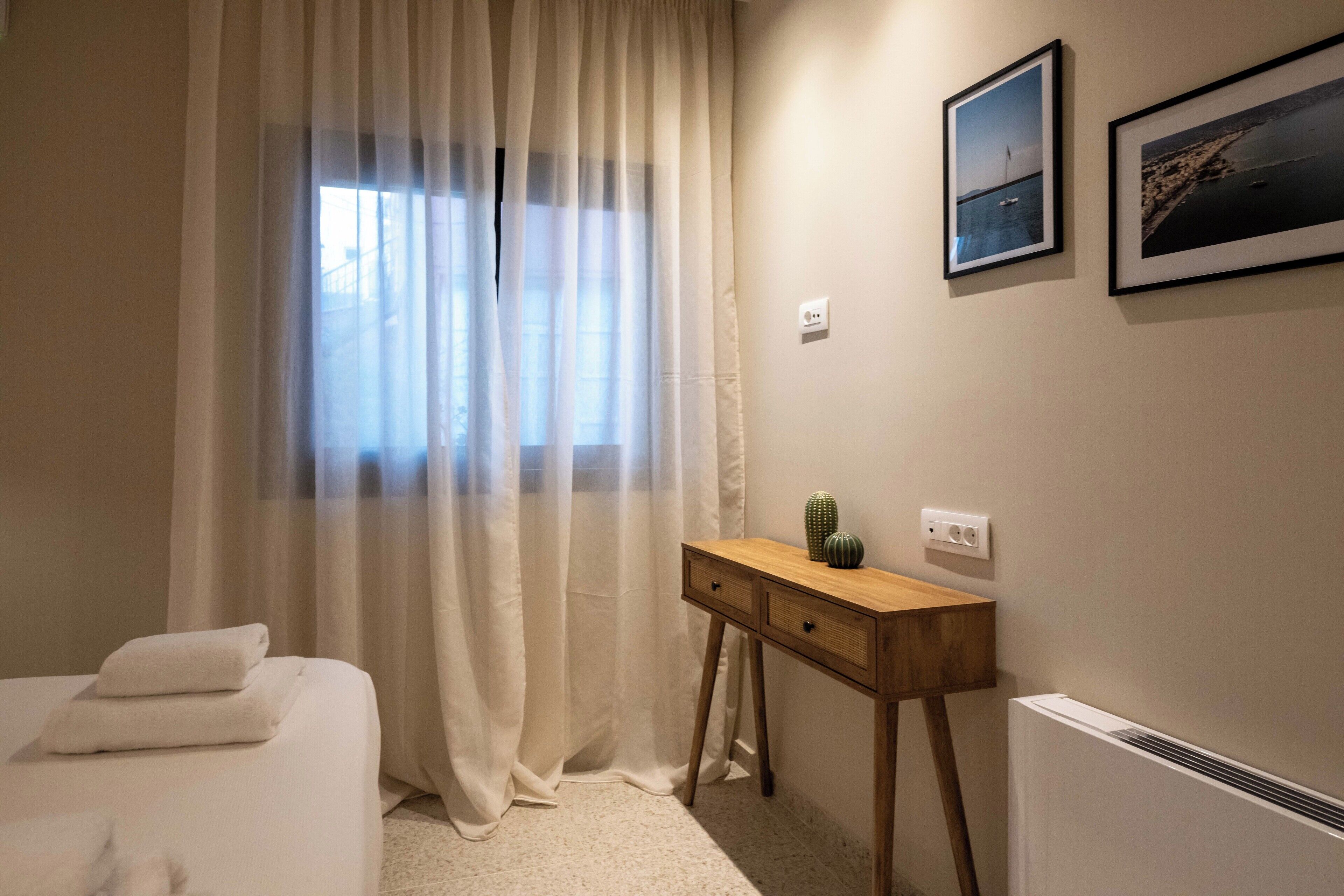A bedroom with a bed, a bedside table with a cactus, a window with sheer curtains, and two framed pictures on the wall.