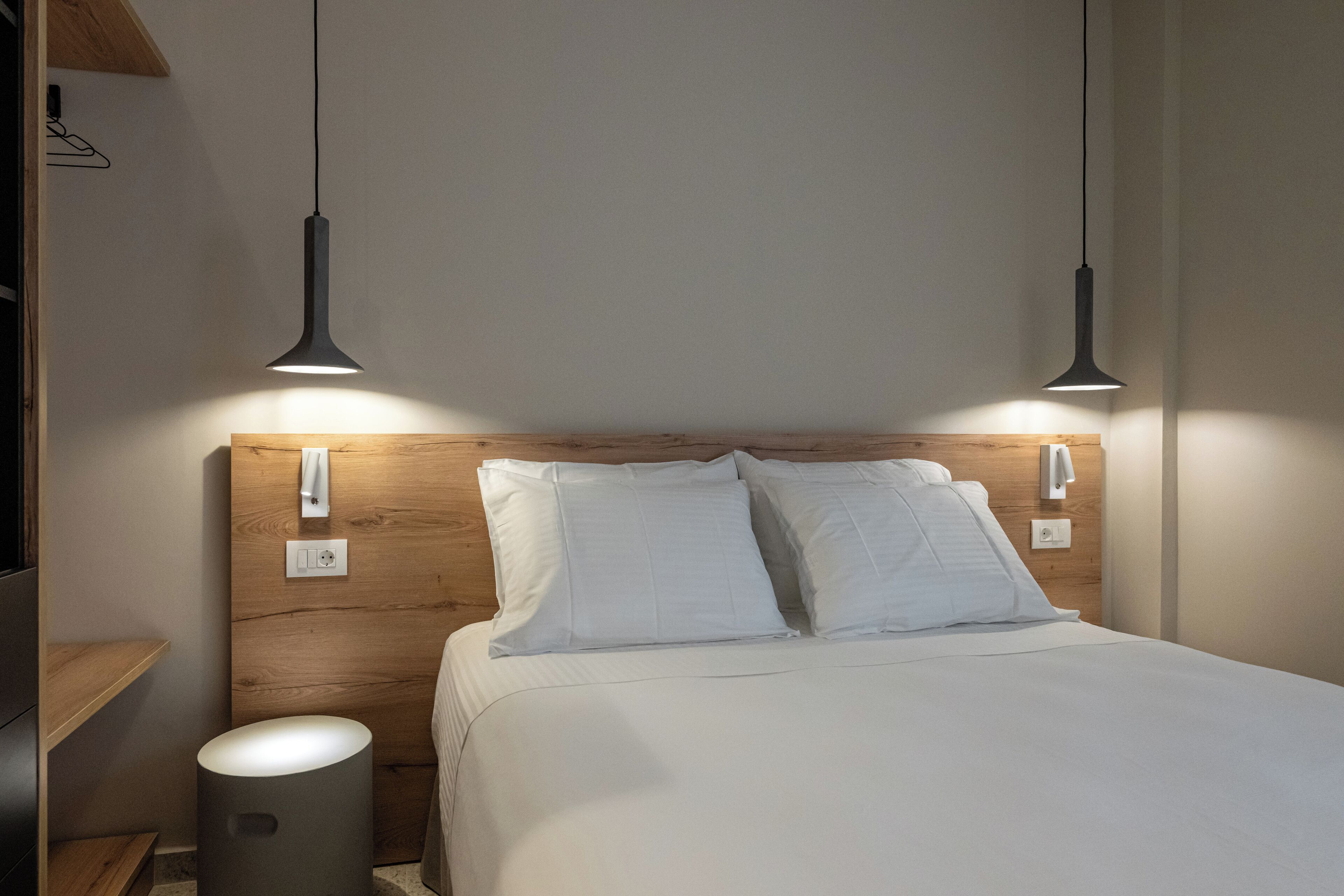 A neatly made bed with white linens, a wooden headboard, and two pendant lights.