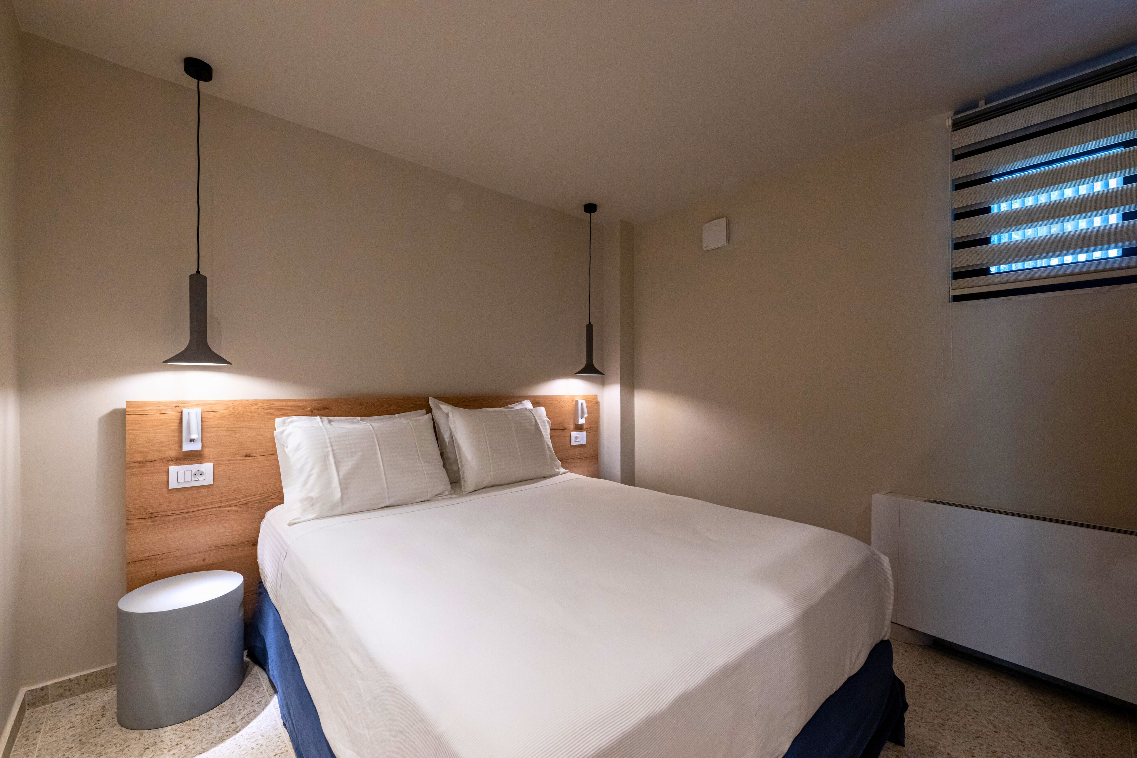 Ett hotellrum med en sänggavel i trä, två sänglampor, en rund papperskorg och ett element.