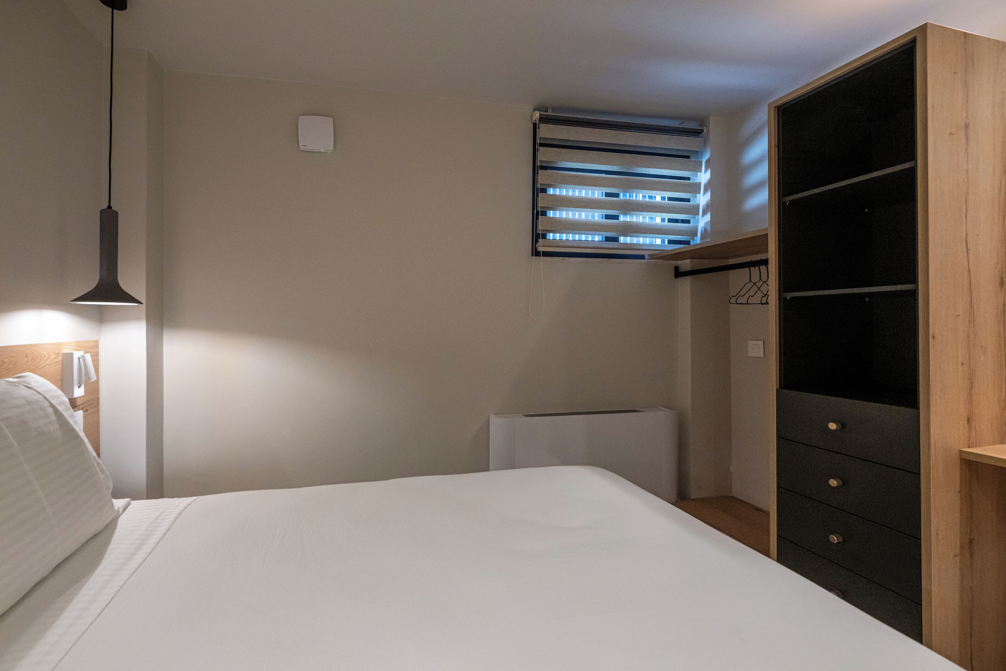 Een slaapkamer met een bed, een hanglamp, een radiator, een raam met zonwering en een kledingkast.