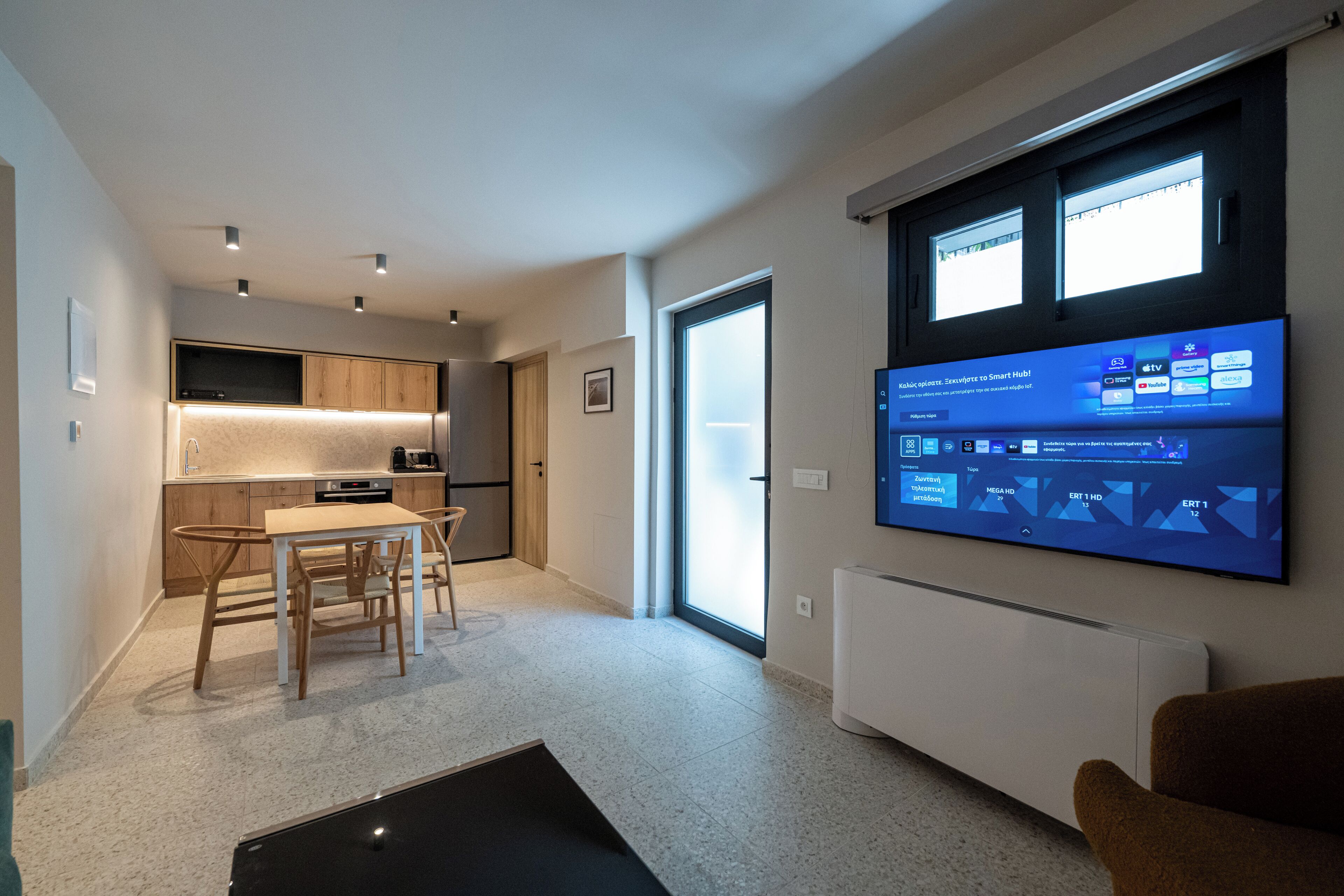 A modern living room with a flat-screen TV mounted on the wall, a dining area with a table and chairs, and a kitchen in the background.