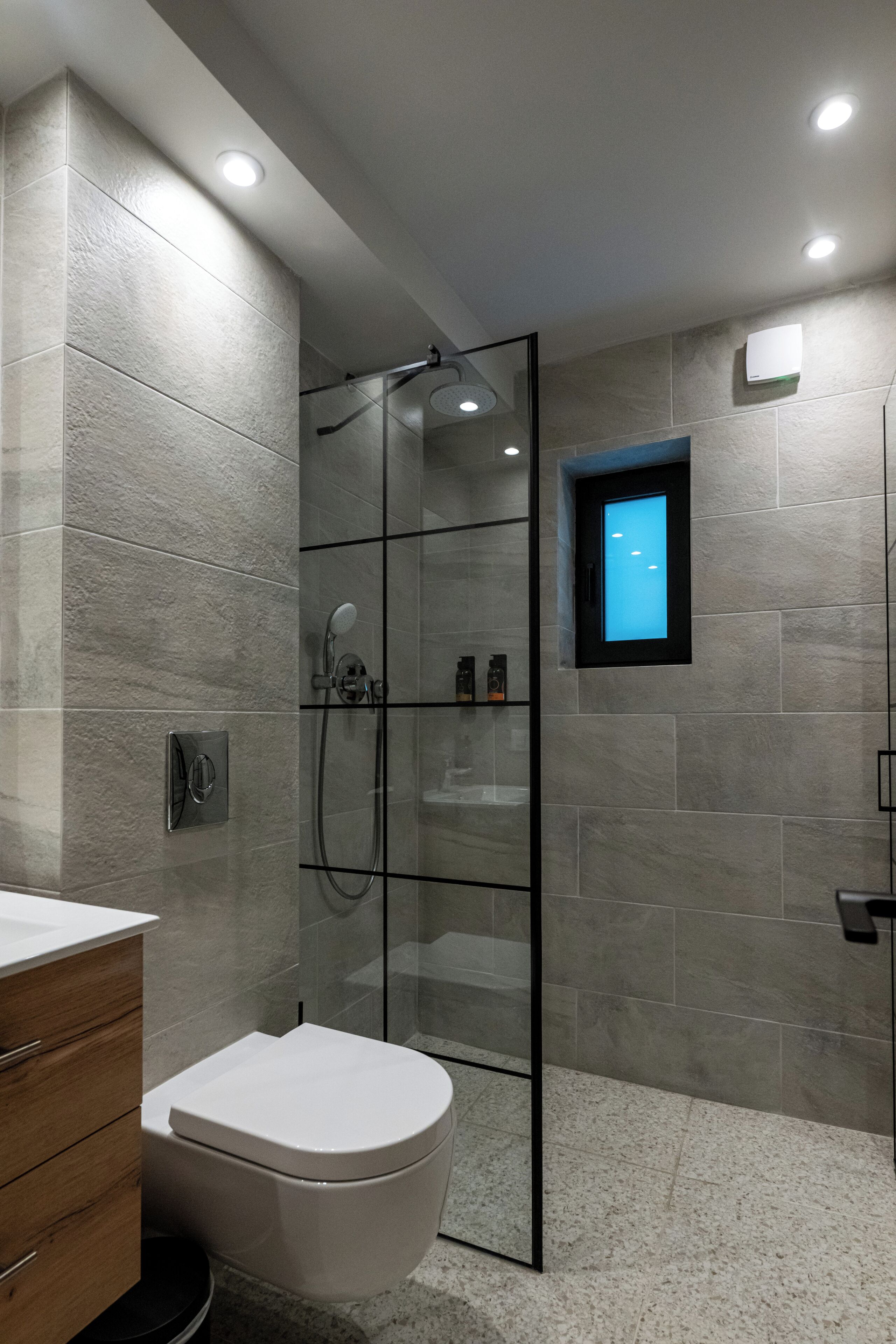 Un baño moderno con ducha envuelta en vidrio, inodoro blanco y una pequeña ventana con luz azul.