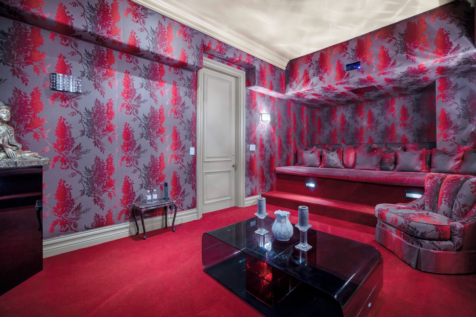 Une pièce avec un tapis rouge, un papier peint à motifs floraux, un canapé et une petite table ornée d’objets décoratifs.