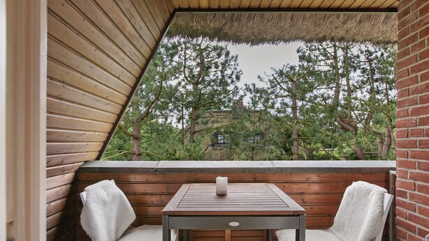 A wooden table with two chairs, a candle, and a view of trees and a building through a triangular window.