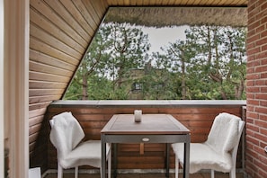 Une table en bois, deux chaises, une bougie et une vue sur des arbres et un bâtiment, le tout visible à travers une fenêtre triangulaire.