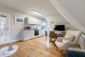 Une cuisine moderne avec des meubles blancs, un coin repas comprenant une table et des chaises, et un espace salon confortable avec un fauteuil et un coussin.