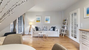 A modern living room with a white sofa, a coffee table, and a lamp. There is a window with curtains, a bookshelf, and a door with glass panels.