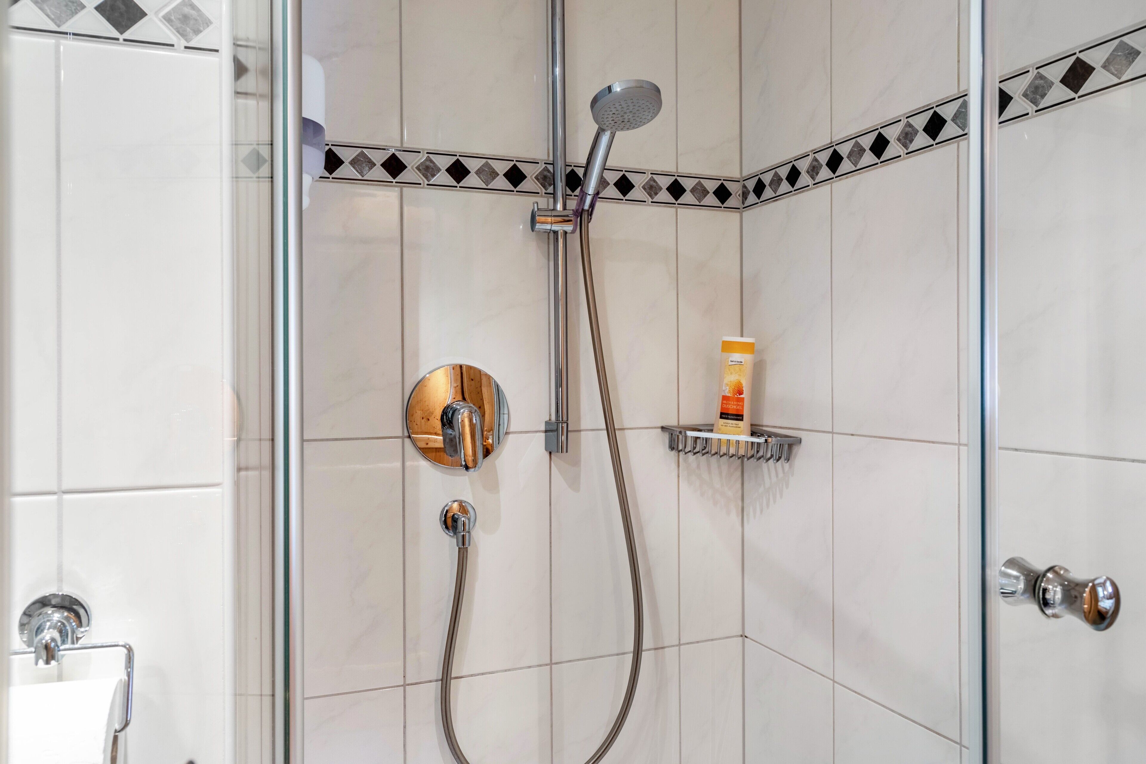 A shower with a handheld showerhead, a wall-mounted faucet, and a soap dispenser on a shelf.