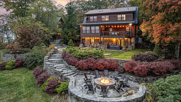 A two-story log cabin with a stone fire pit and outdoor seating area surrounded by landscaped gardens and trees.