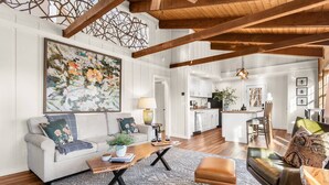A spacious living area with a white sofa, a coffee table, and a leather armchair. A large painting adorns the wall, and a kitchen is visible in the background. - Walk to BE | Hot Tub, Game Room + Ski Access (Banner Elk)