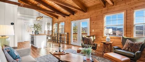 A spacious living room with a wooden ceiling, a dining area, and large windows offering a view of the outdoors.
