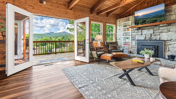 A spacious living room with a stone fireplace, wooden ceiling beams, and a large TV mounted on the wall. - Walk to BE | Hot Tub, Game Room + Ski Access (Banner Elk)
