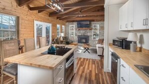 A modern kitchen with wooden beams, a stone fireplace, and a dining area with a view of the outdoors. - Walk to BE | Hot Tub, Game Room + Ski Access (Banner Elk)