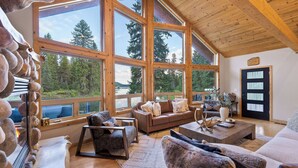 A spacious living room with large windows, a brown leather sofa, and a stone wall. - Riverfront Cabin w/ Hot Tub, Sauna & Fire Pit (Olney)