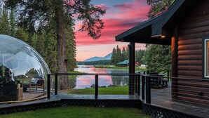 A wooden cabin with a glass dome structure overlooking a lake, surrounded by trees and mountains. - Riverfront Cabin w/ Hot Tub, Sauna & Fire Pit (Olney)