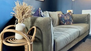 Une pièce avec un canapé vert, un vase blanc contenant des plantes séchées et un fauteuil en osier.