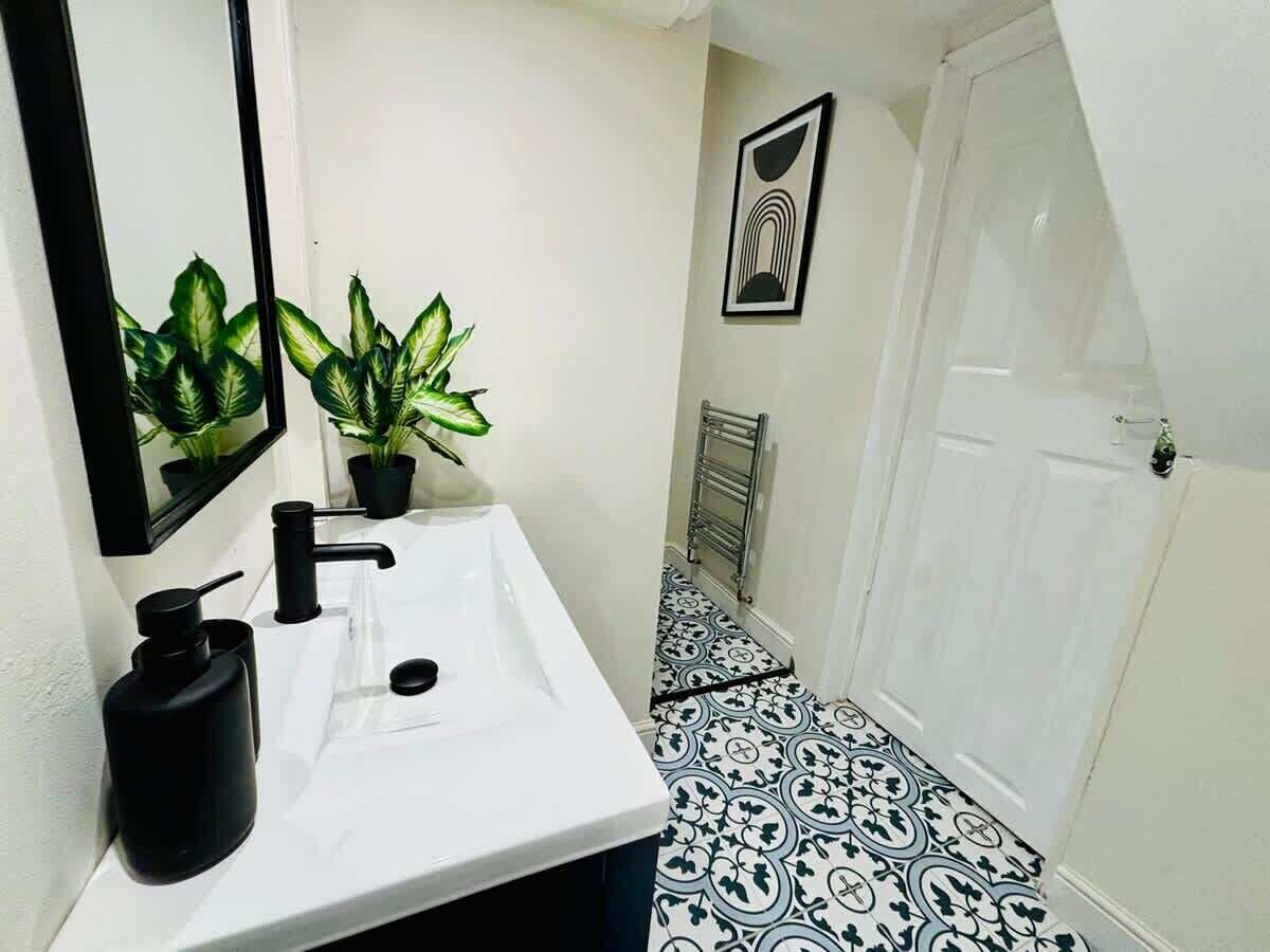 A bathroom with a white sink, black faucet, and black soap dispenser. There are two potted plants on the counter. The floor has a black and white patterned tile design.