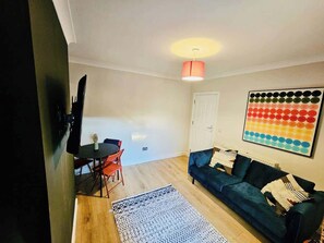 A living room with a dark blue sofa, a patterned rug, a dining table with chairs, and a colorful abstract painting on the wall. - Central Gloucester: Long-Stay Base with Parking (Gloucestershire)