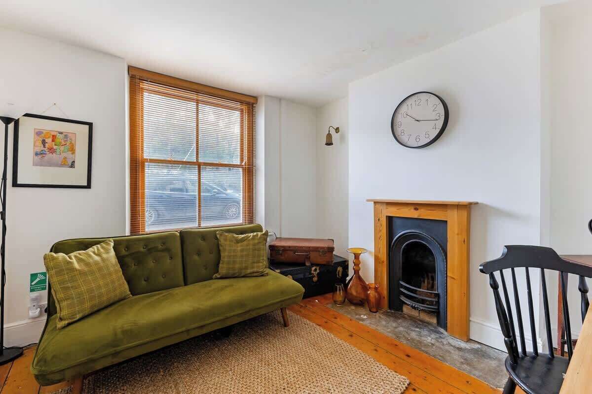 A cozy living room with a green sofa, a fireplace, a clock, and a window with blinds.