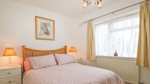 A bedroom with a wooden bed, two bedside tables with lamps, a radiator, and a window with curtains. - Dymchurch Bungalow (Romney Marsh)