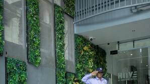 Een beveiligingsbeambte staat bij de ingang van een gebouw met een groene verticale tuin en een glazen deur.