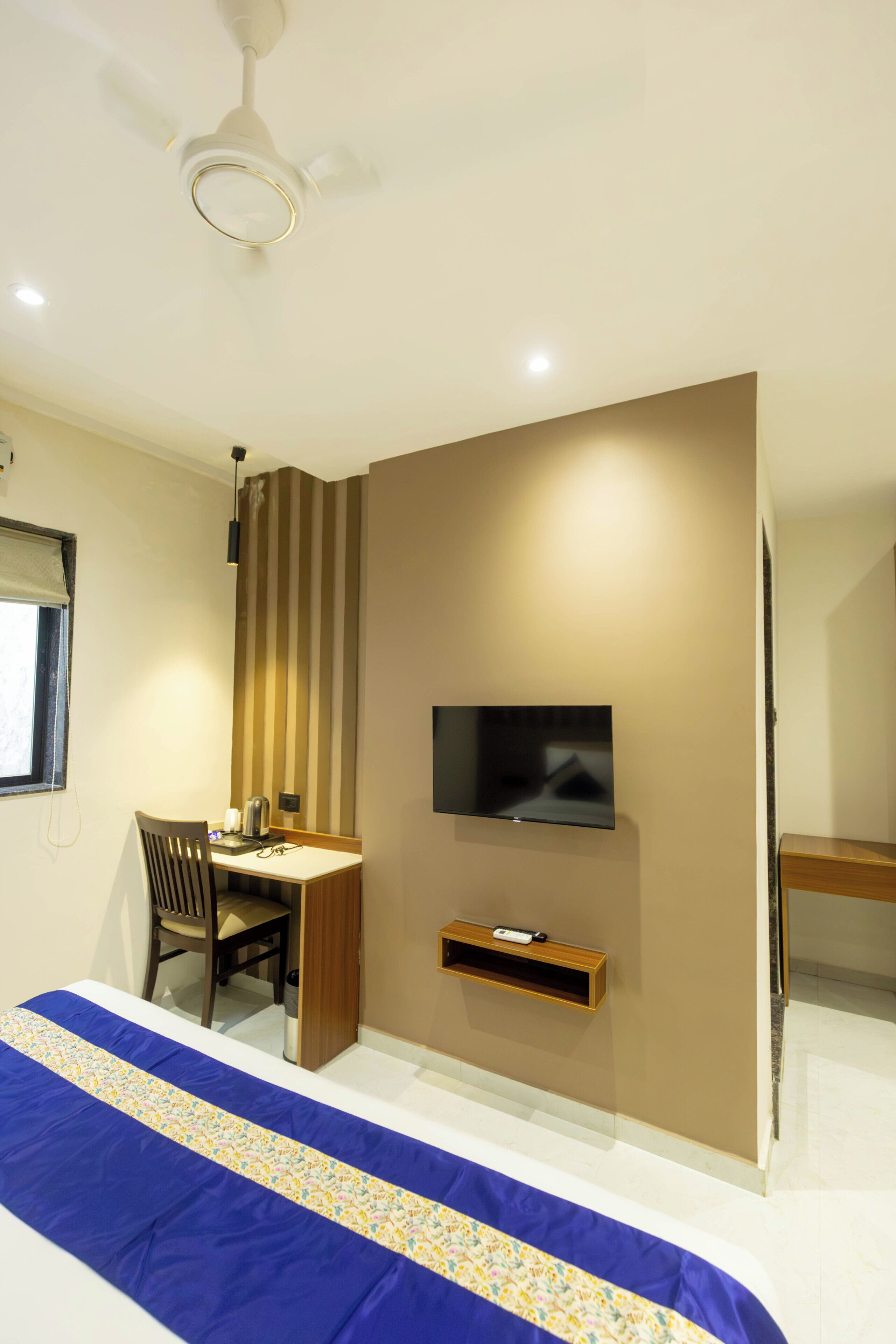 A hotel room with a bed, a desk with a chair, a television mounted on the wall, and a ceiling fan.