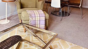 Una stanza con una poltrona verde, un tavolino rotondo con un vaso rosa, una scrivania in legno con una lampada e una sedia bianca.