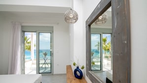 A bedroom with a bed, a wooden bench, a vase with plants, a mirror, and a view of the ocean through a glass door.