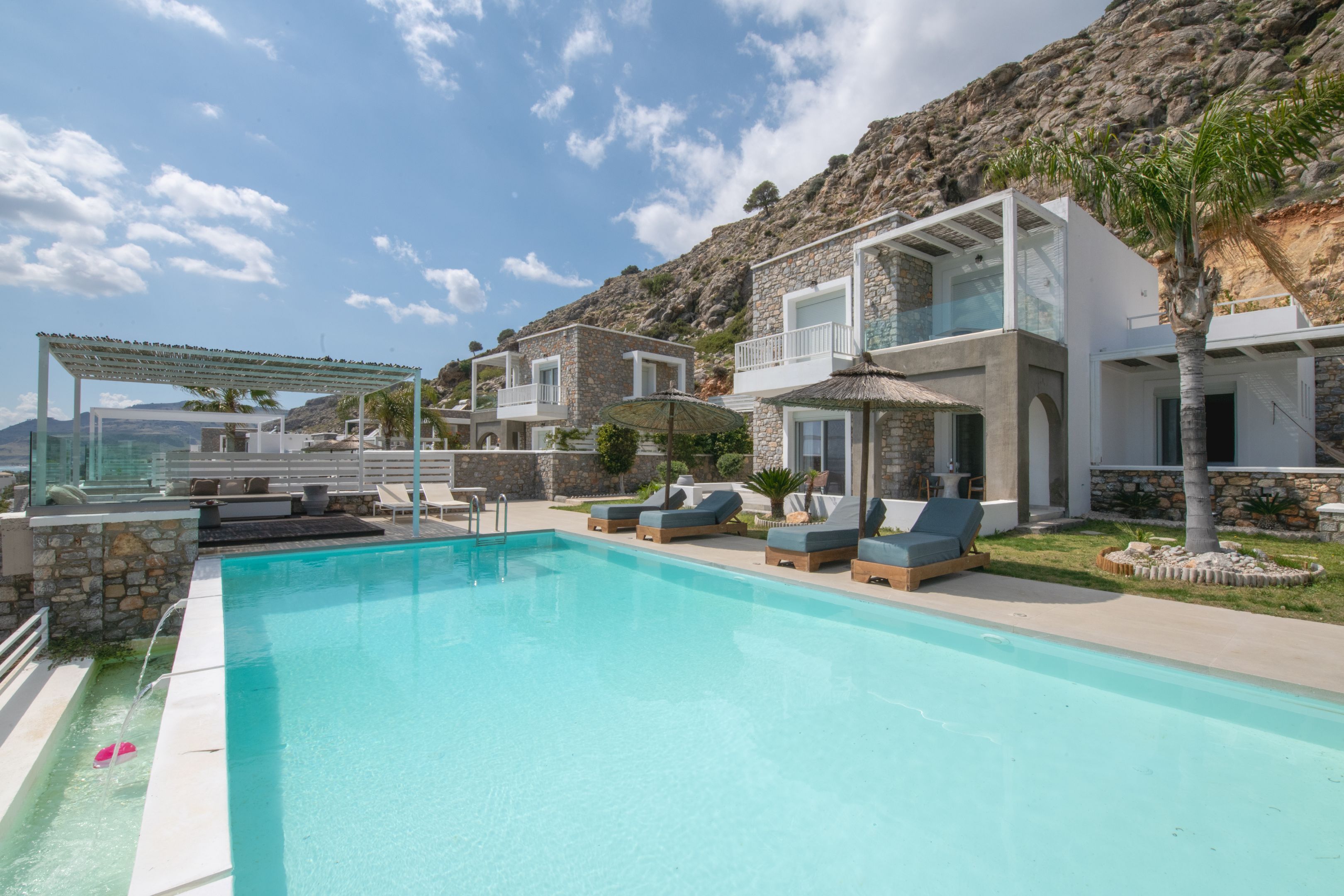 Un área de piscina moderna con sillones de descanso y una pérgola, rodeada de edificios de piedra y un paisaje montañoso.