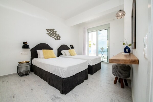 A hotel room with two beds, a small side table, a round ottoman, a wooden shelf, and a view of the outdoors. - Elements Villas (Rhodes)