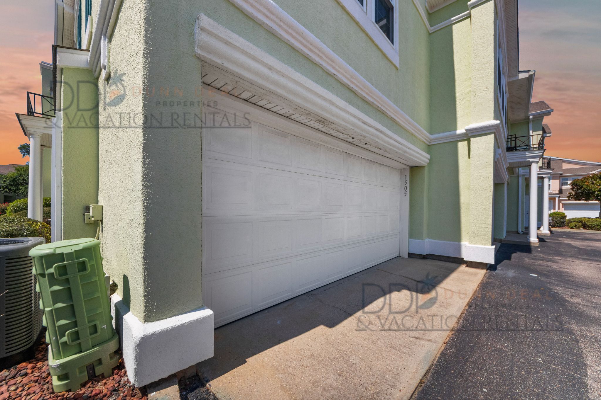 A residential building with a white garage door, a green exterior, and a small air conditioning unit on the left.