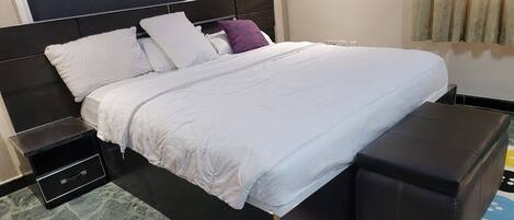 A neatly made bed with white bedding and a black headboard, a small bedside table with a drawer, and a black ottoman with a colorful rug.