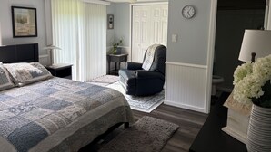 A bedroom with a bed, a chair, a nightstand, a lamp, a clock, and a door leading to another room.