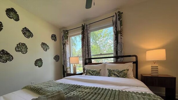Ein ordentlich bezogenes Bett mit grüner Decke und Kissen, eine Wand mit dekorativen Blattmustern und ein Fenster mit Vorhängen.