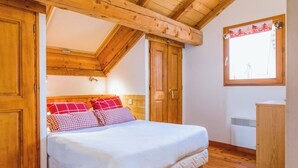 A bedroom with a wooden ceiling, a bed with white bedding, red and checkered pillows, and a window with a curtain.