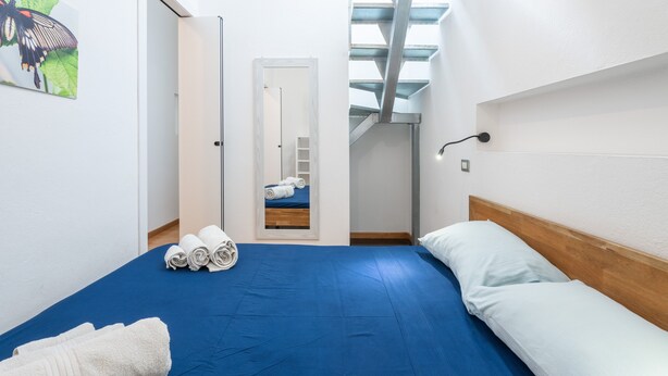 A hotel room with a blue bedspread, white pillows, a wooden headboard, a painting of a butterfly, and a skylight above the bed.