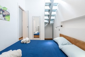 A hotel room with a blue bedspread, white pillows, a wooden headboard, a painting of a butterfly, and a skylight above the bed.