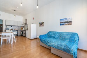 A modern kitchen with white cabinets, a dining table with chairs, a sofa with a blue blanket, and a painting on the wall.