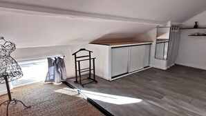 Une pièce avec un plafond en pente, un plancher en bois, une petite table avec une chaise, un radiateur et une fenêtre avec des rideaux.