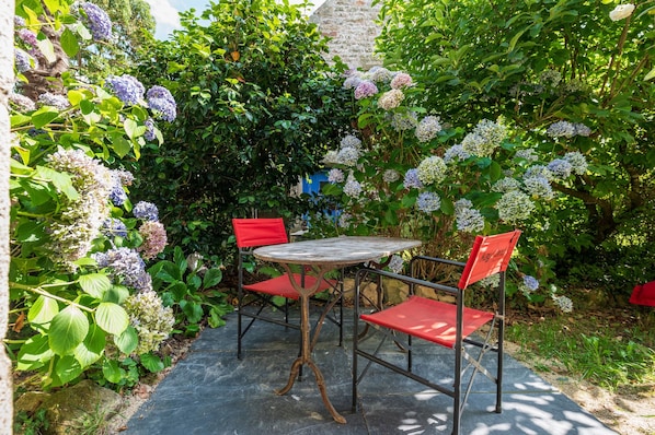 A garden patio with a round table and two red chairs, surrounded by lush greenery and flowering plants.