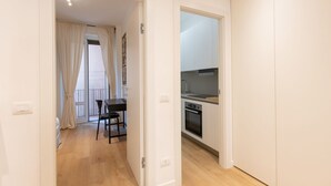 Une porte ouverte donne sur un coin repas avec une table et des chaises, une cuisine équipée d’appareils modernes et un balcon agrémenté de rideaux.